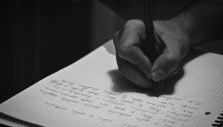 A black and white image of a hand holding a pen and writing in a notebook